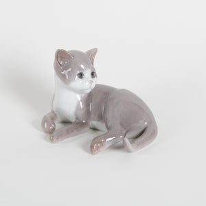 A small glossy ceramic figurine of a lying gray cat with a white chest, photographed on a clean light gray background.