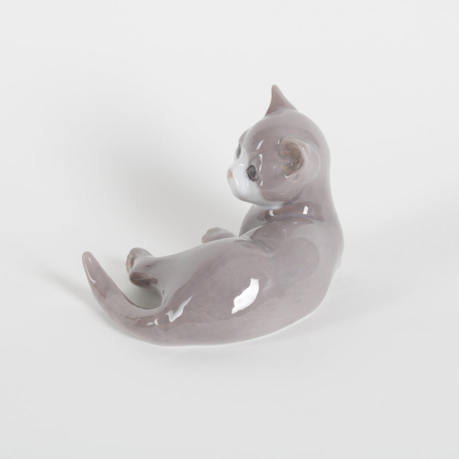 A small glossy ceramic figurine of a lying gray cat with a white chest, photographed on a clean light gray background.