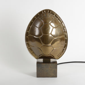 A brass turtle shell lamp sculpture on a dark brass rectangular base, photographed against a plain light gray studio background. A black cord extends from the right side of the base.